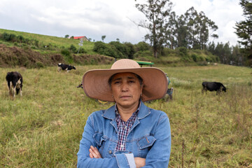 An adult woman in the foreground with a serious expression and crossed arms, standing in front of her herd of cows in a green pasture © Lenin