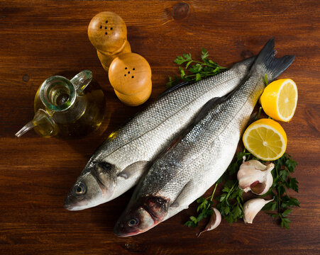 Two raw sea bass fish with lemon, garlic and herbs on wooden table