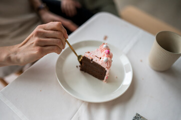 A human's hand is holding a golden spoon stuck in a piece of chocolate birthday cake with pink buttercream and white and pink decorations on a white plate on a white tablecloth next to a creamy cup.