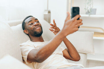 Happy African American Man Using Mobile Phone and Relaxing on Black Sofa at Home Modern Lifestyle...