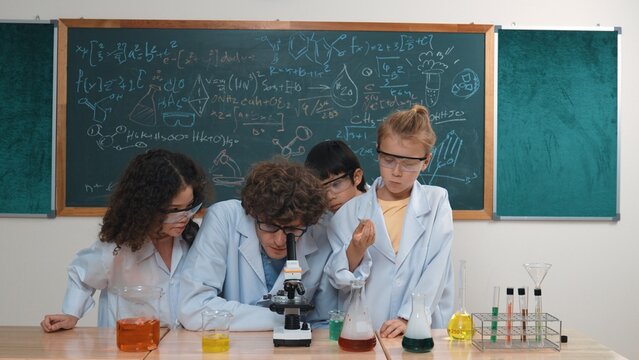 Smart caucasian teacher looking under microscope while diverse children doing experiment at STEM science lesson. Professional happy scientist inspect biochemical example by using microscope. Pedagogy.