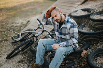 A tired casual young man sits beside a bicycle in the park, embodying relaxation and leisure in the...