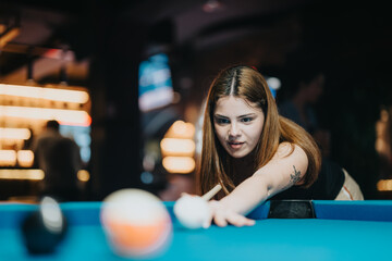 A focused young woman playing billiards, preparing to take a shot in a bustling bar setting,...