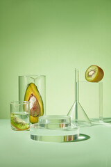 Green background features a stack of round podiums and several laboratory glassware containing avocado and kiwi. Beauty product can be displayed on the glass podium