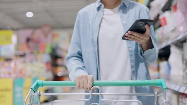 A woman is shopping in a store and is using her cell phone. She is holding a shopping cart and is looking at her phone