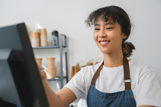 Happy young Asian woman cashier wears an apron and using pos terminal to input orders on coffee shop counter.