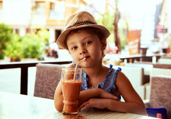 Cute calm thinking kid girl in summer hat drinking tasty juice in street restaurant. Closeup