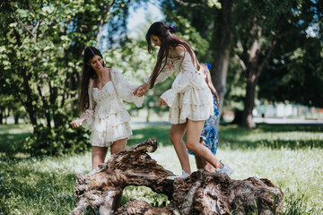 Two young girls in floral dresses have fun balancing on a fallen tree in a lush, sunlit park,...