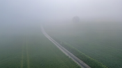 An aerial view shows a road passing through a fogcovered field, creating a mysterious atmosphere. The landscape includes a plain with tall grass and a hazy horizon under the sky