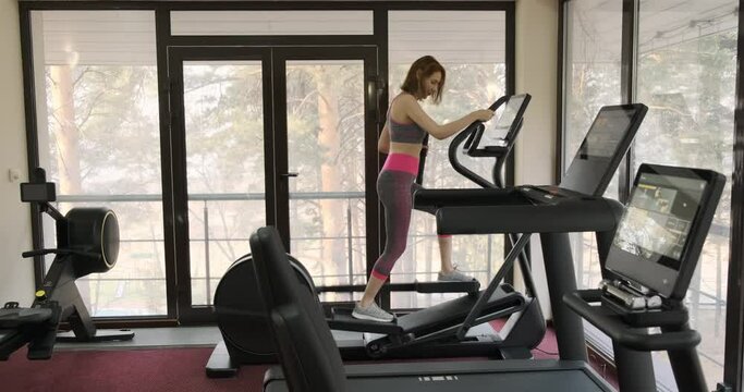 Middle aged woman working on stepper and looking at fitness bracelet while checking her activity and heart rate, gym workout. Girl is working out at gym. Young woman in sportswear out at sport hall