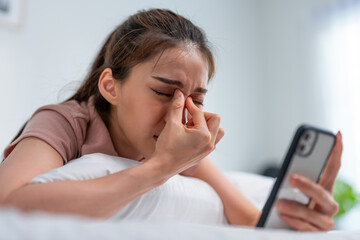 Exhausted Asian young woman hurt eye while using mobile phone on bed.