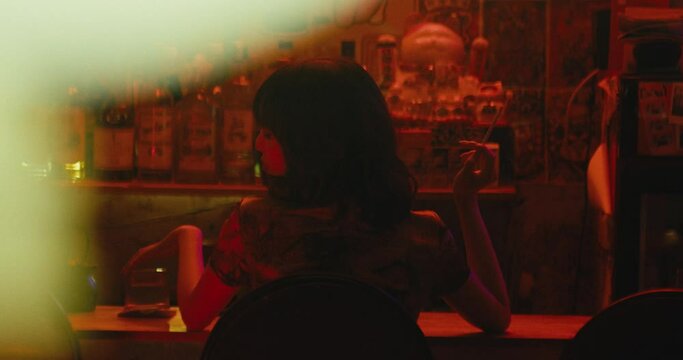 sad woman sit alone in cafe bar club while drinking a cocktail and smocking a cigarette from view