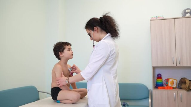 female doctor in a white coat uses a stethoscope to examine a young boy sitting on an examination table in a pediatric clinic. The room is equipped with medical and children's toys