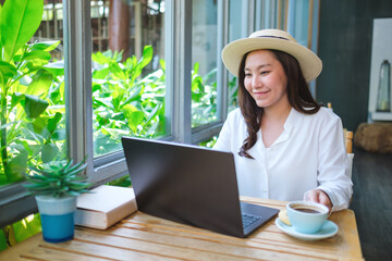 Portrait image of a young woman with hat drinking coffee while working on laptop computer in cafe