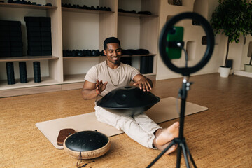 Cheerful black male blogger practicing native music traditional drumming instrument sitting on floor in front of mobile phone with ring light. Concept of sound therapy, mindfulness, tranquility.