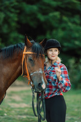 Happy blonde with horse in forest. Woman and a horse walking through the field during the day. Dressed in a plaid shirt and black leggings.