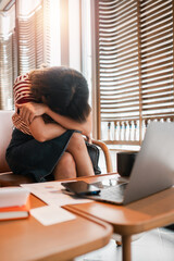 A woman is sitting on a chair with her head on her hands. She is crying and has a laptop on the...