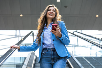 Smiling young business woman wearing suit standing on urban escalator using applications on cell...