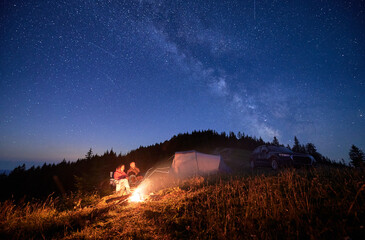Night camping in the mountains under starry sky full of stars. Travelling couple having a rest at campfire, tourist tent and off-road vehicle in the evening on mountain lawn and stargazing together.