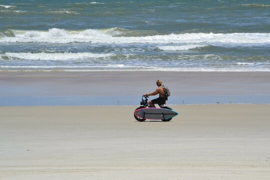 A surfer glides along on his electric bike, surfboard in tow, across the sandy shores of Ponce Inlet, Florida, while the waves break tumultuously against the horizon.