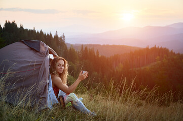 Woman traveller camping outdoors at sunset. Beautiful female sitting in tent's entrance on grass...