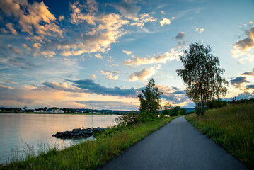 Promenade or a bicycle track on the bank of the Danube river at sunset with green trees and a...