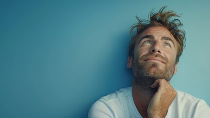 Obraz premium A bearded man in a white shirt leans against a blue wall, touching his chin