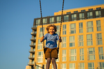 Excited kid swinging on chain swing on city kids playground. Swing ride. Cute child having fun on a swing on summer sky background. Blonde little boy swings at kid playground. Child swinging high.