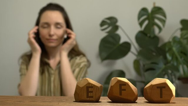 Letters EFT writen on irregular wooden blocks. Female tapping side of the eye  (SE) meridian point in blurred background. Emotion-focused therapy treatment concept.