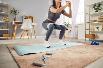 Obraz premium Young sporty woman doing stretching exercises with rubber band standing in living room at home. Happy athletic girl doing domestic workout. Sport, fitness and home training concept.