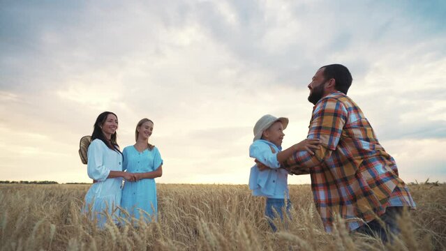 Happy family dad, mom, son and daughter walk on wheat field at sunset in summer. Father throws son in air, plays with him, mother and teen girl smiling. Relaxed people having good weekend together.