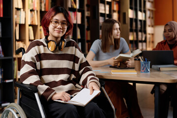 Red-haired Asian girl with disability sitting in wheelchair in modern college library looking at...