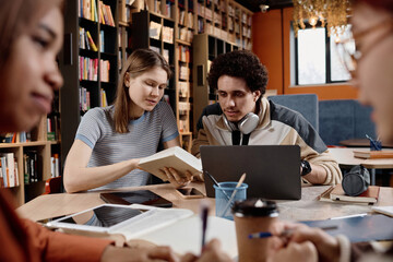 Selective focus shot of young male and female college students sitting in library doing school project using book and laptop - Powered by Adobe