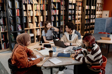 High angle view of four ethnically diverse male and female college students sitting in modern university library doing homework