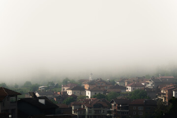 Bulgaria - 05 22 2024: Fog over a big village with small white church in the center in the background being covered in fog.