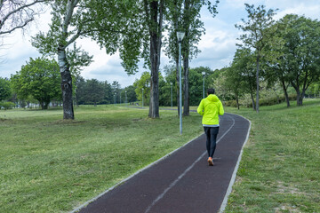 A runner in a bright yellow jacket and black pants is seen from behind, jogging on a well-maintained park path surrounded by lush green grass and trees under a cloudy sky.