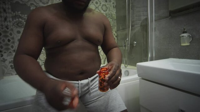 Obese Black man sitting on edge of bathtub and taking weight loss medications in morning at home