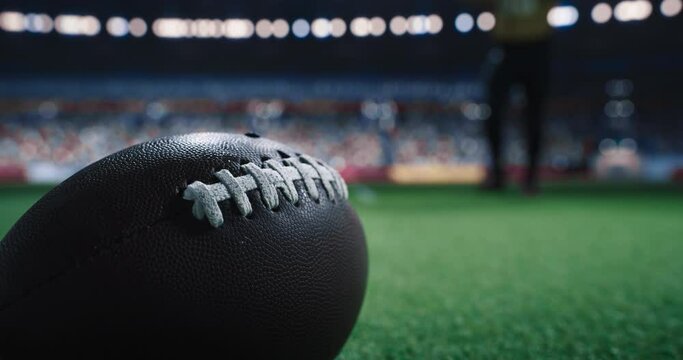 Close Up View of a Brown American Football Pigskin on a Green Pitch, Illuminated by Stadium Lights with Cheering Crowd of Fans in the Background. Fit Athlete Preparing for a Championship Game