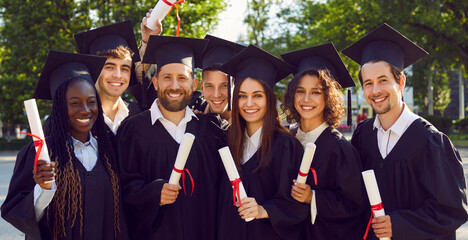 Portrait of a group of smiling happy multiracial international graduates students standing in a row in a university graduate gown and holding diploma. Education and graduation concept. Banner.