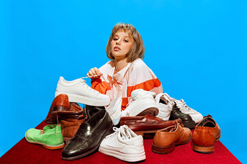 Fashion and Footwear. Stylish young woman sitting at table with various shoes forming pyramid on blue background. Shoe sore advertisement. Concept of creative photography, pop art style, imagination