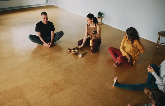 Smiling female yoga instructor using a singing bowl and chime to begin a meditation class of students sitting together on the floor of her studio