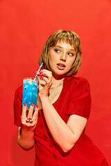 Vivid Refreshment. Beautiful young woman with makeup, in dress drinking bright blue cocktail with fake eyes inside against red background. Concept of creative photography, pop art style, imagination