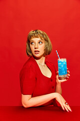 Beautiful young woman in red outfit, holding striking blue beverage, unusual cocktail against red...