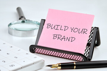Conceptual image with word BUILD YOUR BRAND on a pink sticker on a black stand on a white background