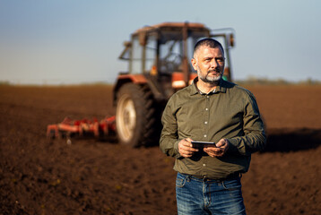 Portrait of satisfied mature farmer standing in field with tablet supervises the cultivation of...
