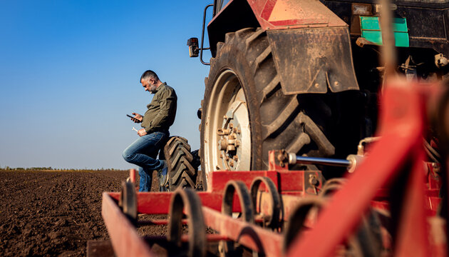 Farmer using smart phone while resting after cultivation of soil leaning on tractor tire.
