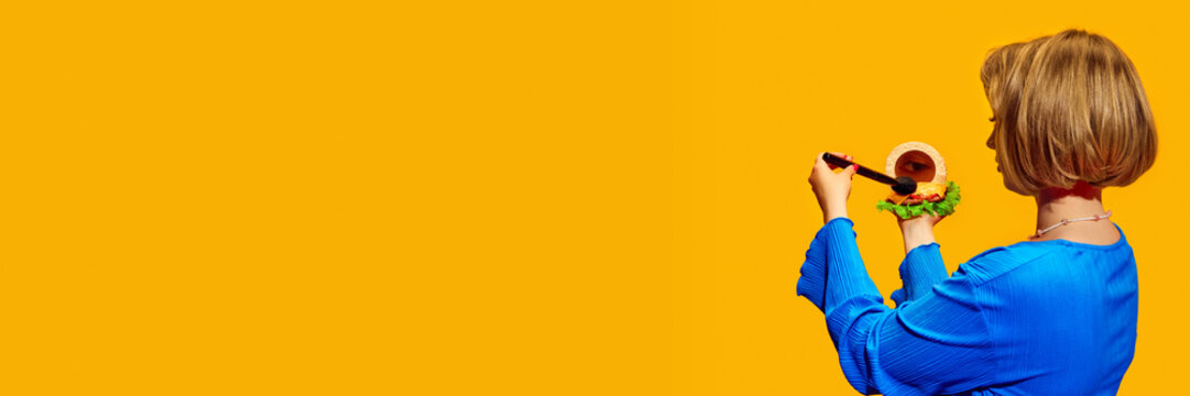 Creative humorous woman applying makeup with brush from blushed with burger element against bright yellow background. Concept of creative photography, pop art style, imagination. Banner for ad