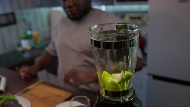 Overweight African American man putting green apple slices into kitchen blender while making healthy smoothie at home