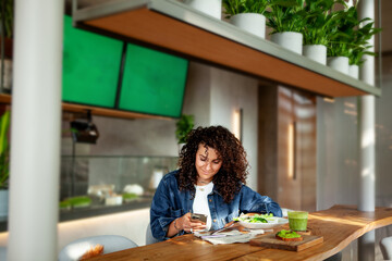 Woman with curly hair seats at vegetarian cafe or restaurant with green smoothie and plant meal on...