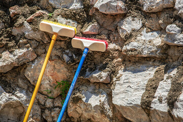 Two colorful cleaning floor brushes with long handle leaning on the stone wall background, out door...